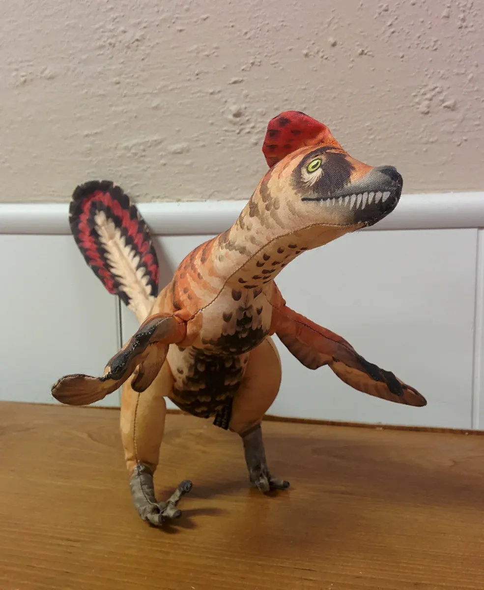 Feathered poseable Velociraptor plush toy by Rebecca Groom, standing on a wood floor with a red-and-black crest and wing feathers