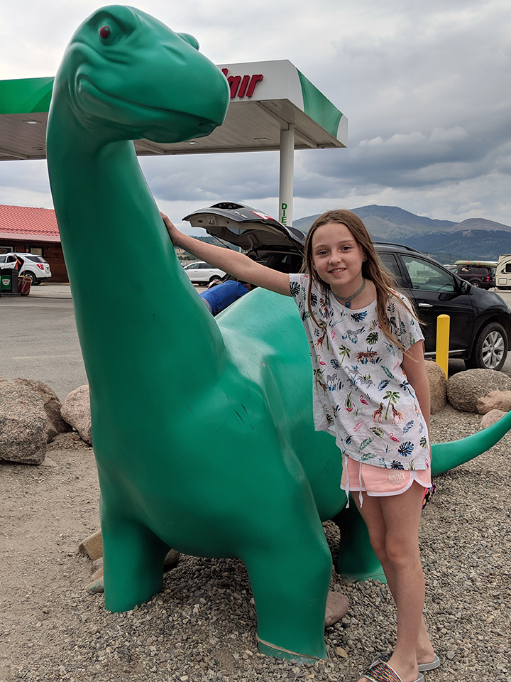 A young girl stands next to a green sauropod dinosaur at a Sinclair gas station